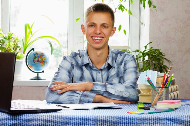 A blue-eyed young man, dressed in a plaid shirt, sits at his workplace and smiles . Pandemic. Education concept.