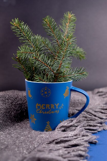 Blue festive Xmas mug with fir branches on grey knitted plaid with fringe on black wall. Cozy home and hugge winter, Christmas or New Year atmosphere. Vertical orientation. Selective focus.