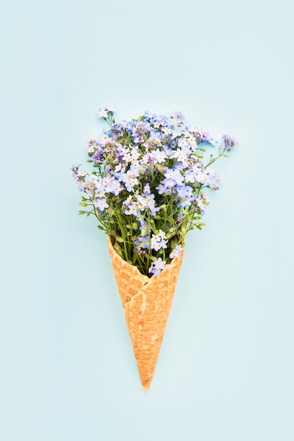 Blue forgetmenot flowers in waffle ice cream cone on light blue background summer concept top view