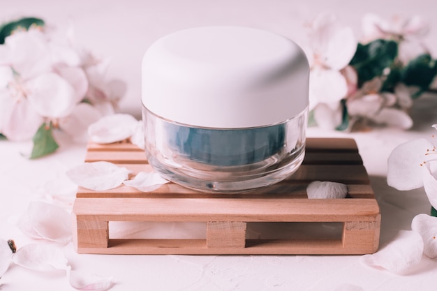 Blue glass jar of cream on wooden podium in form of pallet, on light plaster surface with apple flowers