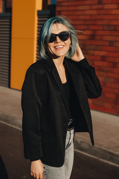 Blue haired businesswoman posing outside in the street wearing eyeglasses near a brick stone wall and smile