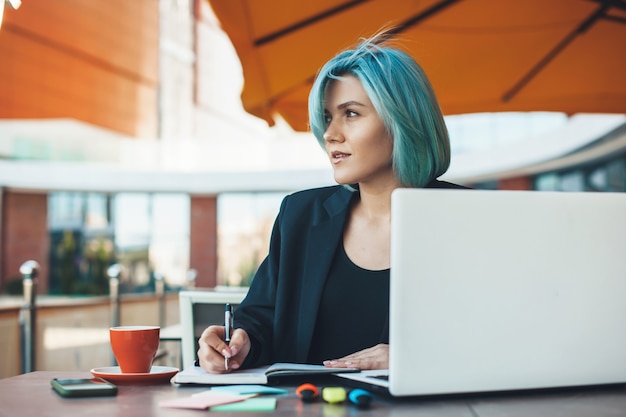 Imprenditore caucasico dai capelli blu seduto in terrazza e lavorando con alcuni documenti al pc