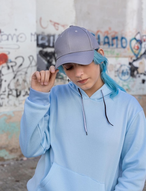 Blue haired Teenage girl in blue hoodie and baseball cap with closed eyes touching her cap Blue haired teen girl stays outdoors against graffiti wall Clothing and cap mockup