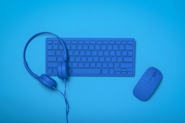 Blue headphones on a blue keyboard and a blue mouse on a blue surface. Monochrome image of office accessories.