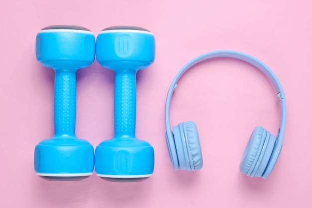 Blue headphones, plastic dumbbells on pink pastel background.