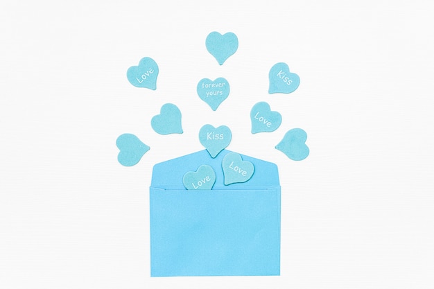 Фото Синие сердца с текстом любовь, поцелуй, навсегда вылетают из синего конверта на белом фоне.