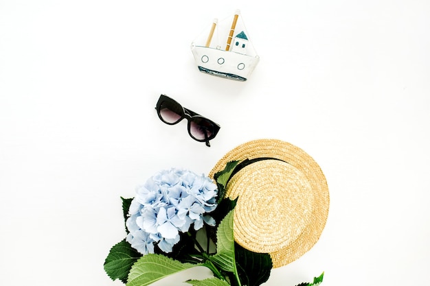 Blue hydrangea flower, straw hat, sunglasses, toy boat on white surface