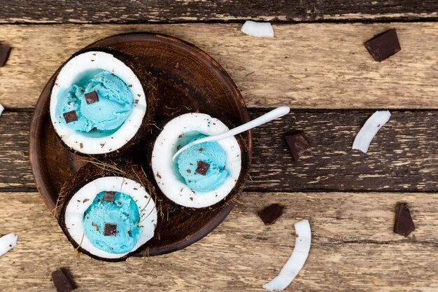 Blue ice cream with chocolate in coconut bowl on wooden background. Summer food. Top view. Copy space.