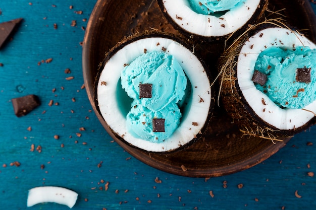 Blue ice cream with chocolate in coconut bowl on wooden background. Summer food. Top view. Copy space.