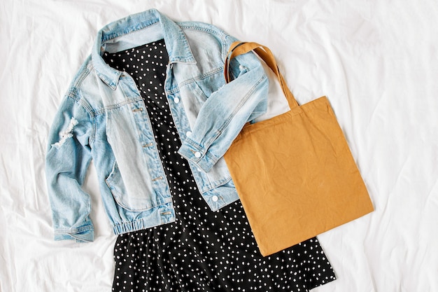 Blue jean jacket and black dress with tote bag on white bed.\
women\'s stylish autumn or spring outfit. trendy clothes. flat lay,\
top view.