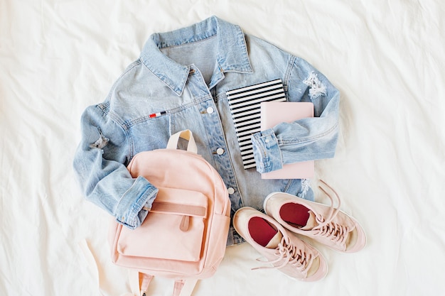 Blue jean jacket pale pink with backpack, book and sneakers on white. Women's stylish autumn or spring outfit. Trendy clothes for college. Back to school concept.  Flat lay, top view.