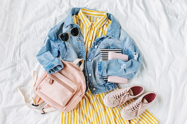 Blue jean jacket and Yellow dress  with backpack, book and sneakers on white. Women's stylish autumn or spring outfit. Trendy clothes for college. Back to school concept.  Flat lay, top view.