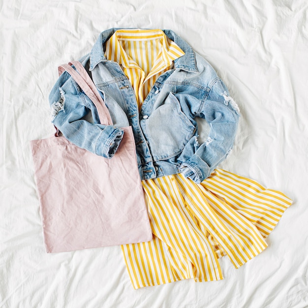 Blue jean jacket and yellow dress with eco bag on white bed.
women's stylish autumn or spring outfit. trendy clothes with eco
bag mockup. fashion concept. flat lay, top view.