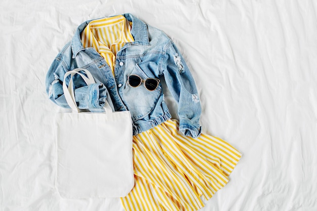 Blue jean jacket and yellow dress with eco bag on white bed.\
women\'s stylish autumn or spring outfit. trendy clothes with white\
eco bag mockup. fashion concept. flat lay, top view.