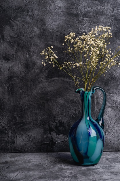 Синяя кувшинная ваза с сыпучими гипсофиловыми белыми цветами на темной фактурной каменной стене, угол обзора