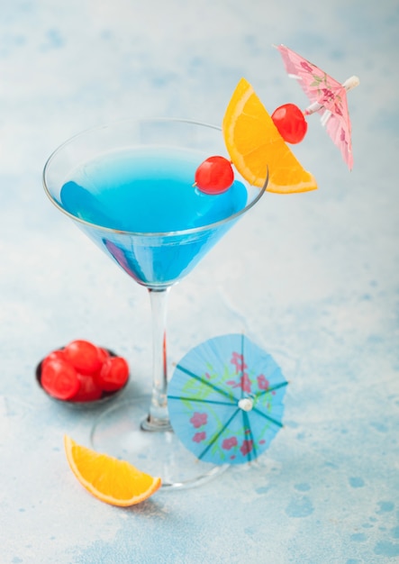 Blue lagoon summer cocktail in martini glass with sweet cocktail cherries and orange slice with umbrella on blue table background.