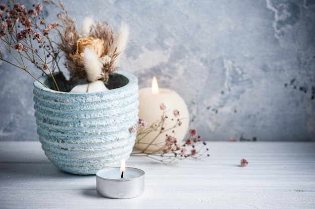 Blue lit candle and dry flowers in pot on white wooden table.