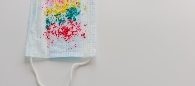 Blue medical mask, sprinkled with multicolored holi paints on white background, close-up. Concept of celebrating the spring festival of colors during coronavirus pandemic. Copy space.