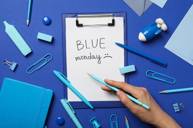 Blue Monday text stationery and hand with pencil on blue background top view