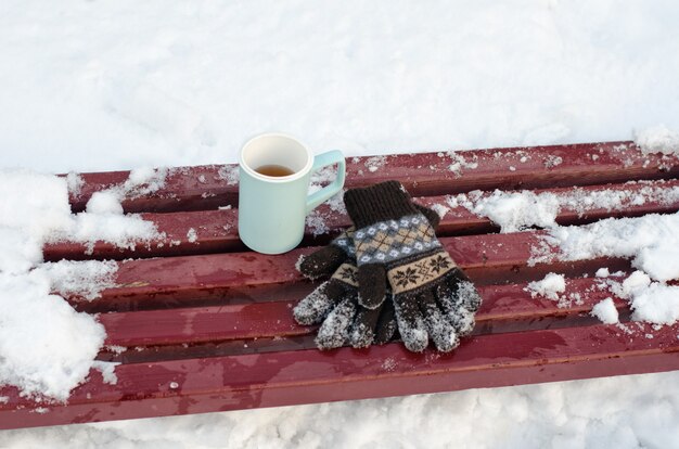 Blue mug tea and women's knitted gloves on a bench in the winter, snow in the background. Close-up