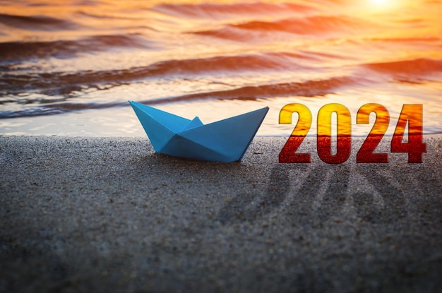 Blue paper boat and year 2024 on sandy beachAgainst background sunset