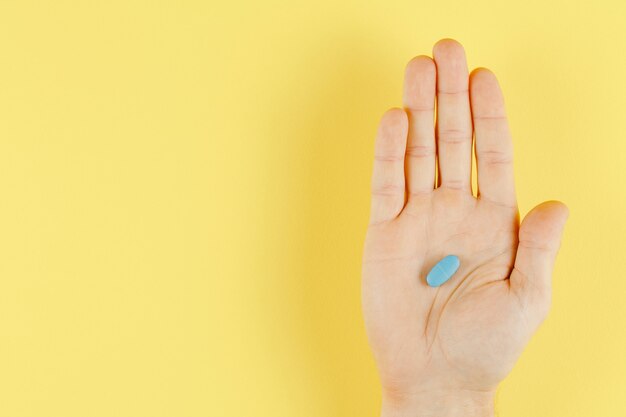 Blue pill in hand on yellow wall top view, flat lay and lot of free space, medical wall