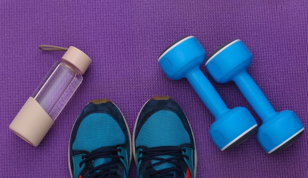 Blue plastic dumbbells, sport shoes, water bottle on a purple background. Top view. Flat lay