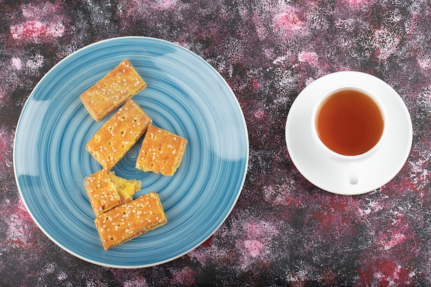 Blue plate of biscuits with sesame seeds and cup of tea on purple table.