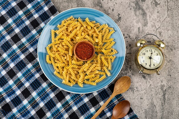 Blue plate of raw fusilli and alarm clock on stone table.