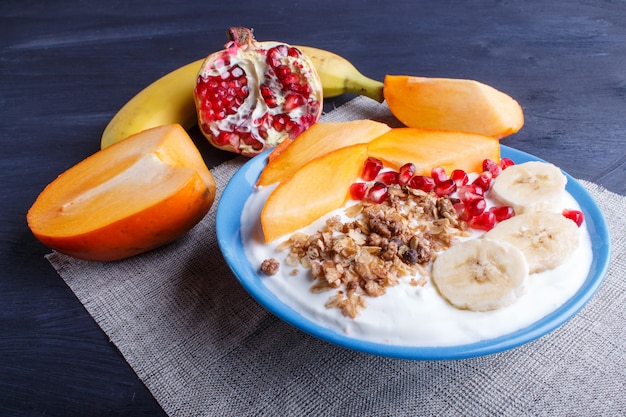 A blue plate with greek yogurt, granola, persimmon, banana, pomegranate