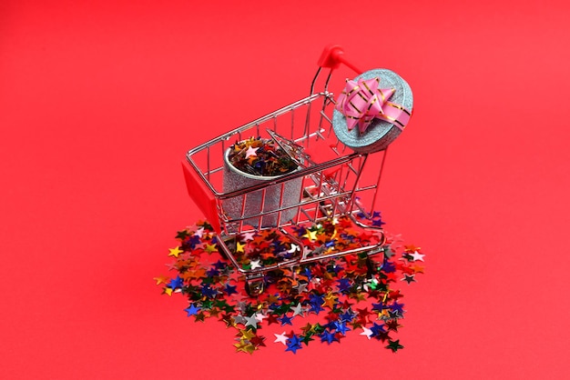 Blue present box with pink bow in a shopping cart and confetti on a red background