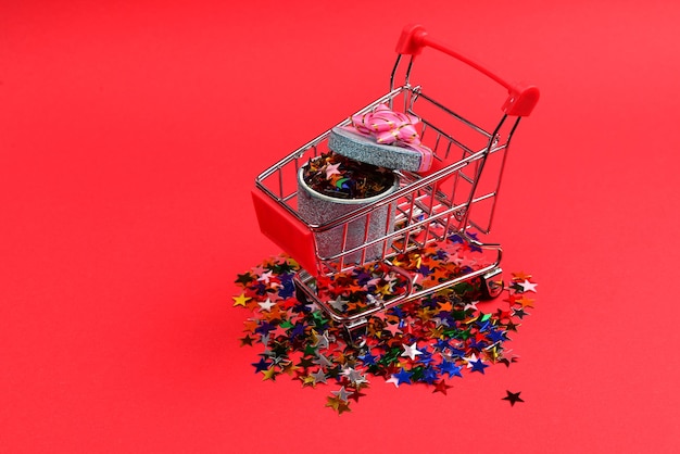 Blue present box with pink bow in a shopping cart and confetti on a red background