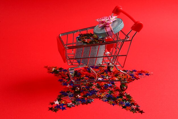 Blue present box with pink bow in a shopping cart and confetti on a red background