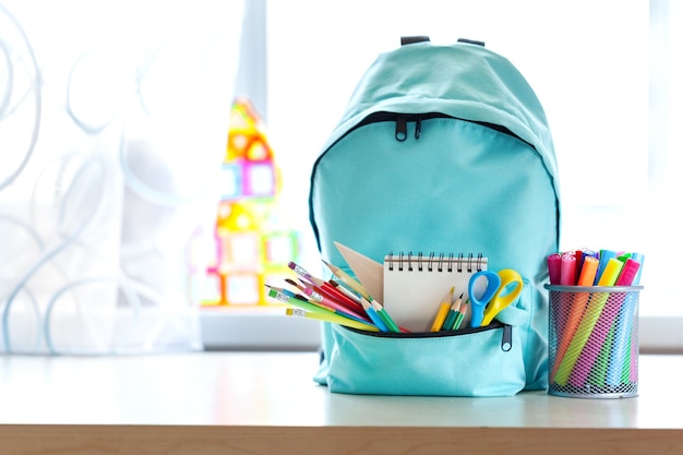 Zaino scuola blu con materiale scolastico sul tavolo sopra l'interno della stanza dei bambini nella giornata di sole