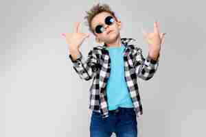 Photo blue shirt and jeans stands on a gray background. the boy is wearing round glasses. redhead boy shows a rocker goat