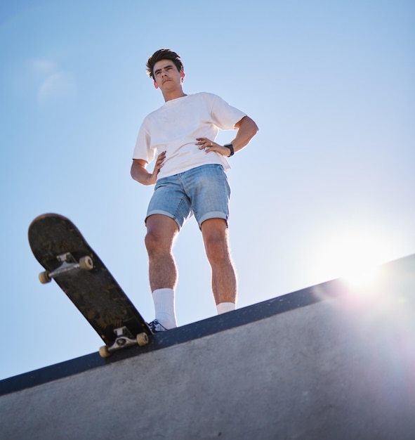 Blue sky skateboard and young man from below on skate ramp ready for trick Fitness street fashion and urban sports a skater in skatepark and summer sun for adventure freedom and extreme sport