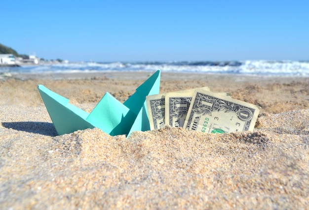 Barca di carta blu piccola e banconote da tre dollari a metà nella sabbia sulla spiaggia vicino al mare in estate soleggiata da