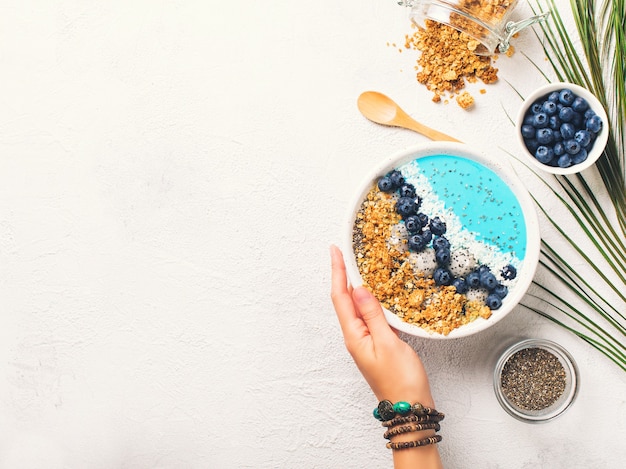 Blue smothie in bowl with granola and berries, holding hand 