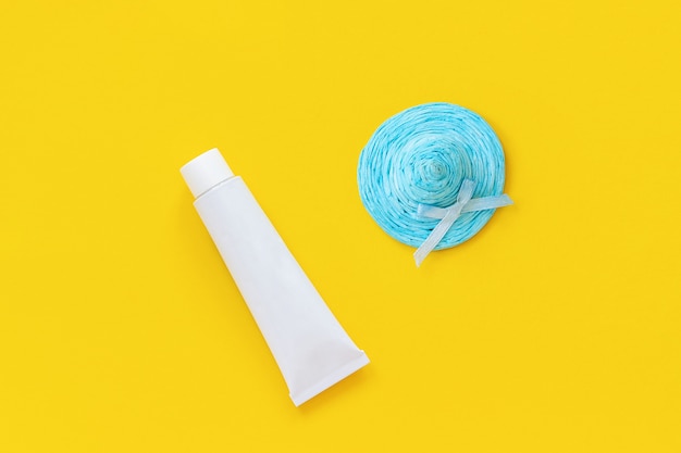 Blue straw hat and white tube of sunscreen on yellow paper background