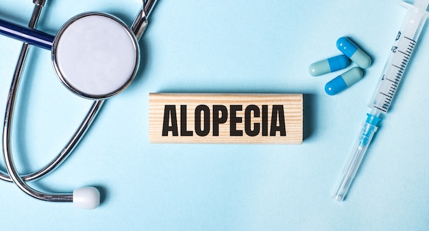 На синей поверхности стетоскоп, шприц, таблетки и деревянный брусок с надписью ALOPECIA.