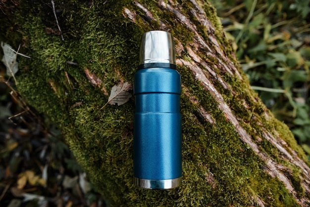 Blue vacuum thermos outdoors. Tree background with green moss. Top view. Concept  hot drink, hike