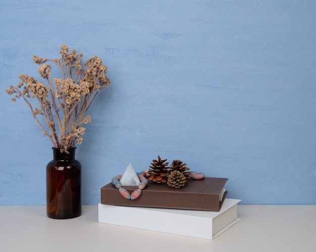 デザインやテキスト、白と茶色の本、ガラスの瓶にドライフラワー、松ぼっくり、木製のテーブルの上に座っているアンティークの石のための青い壁のコピースペース