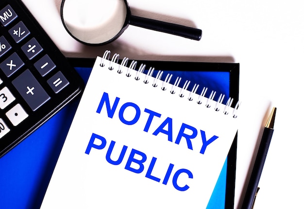 파란색 벽, 계산기, 돋보기 및 펜 근처에 NOTARY PUBLIC이라는 비문이있는 노트북.