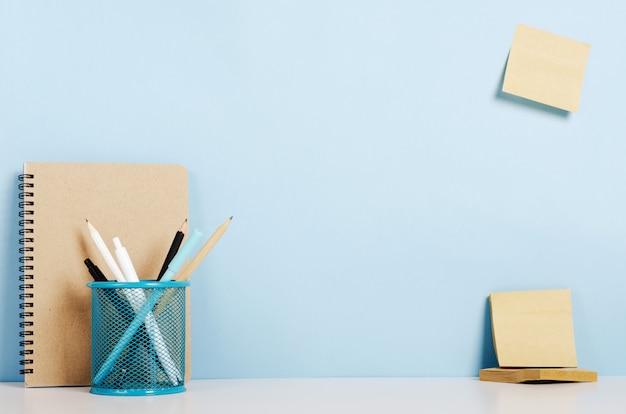 Blue, white and black pencils, pens in the stand, craft notebook on a white table, stickers on the pigeon wall, office desk.