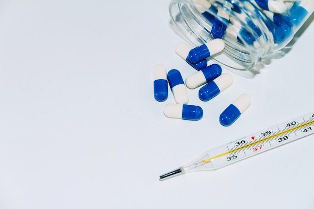 Blue and white capsules spilling out of bottle and thermometer for temperature measurement closeup on a white background Top view with copy space Medicine concept