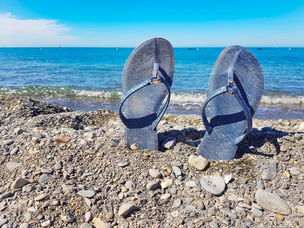 Blue women's beach shoes standing in the pebbles against the background of the sea and the blue sky Closeup a place for text the concept of a beach holiday by the sea