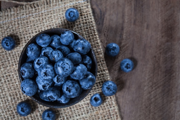 Photo blueberries in a brown ceramic bowl placed on sack and wooden table, taken on top view