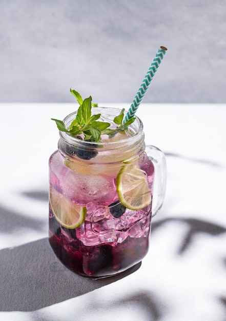 Blueberry mojito or lemonade with lime ice and mint in a glass on a light background with berries fruit and shadows The concept of a summer refreshing drink