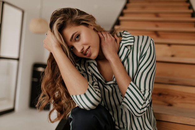 Blueeyed blonde curly woman smiles gently touches her hair looks into camera and sits on wooden