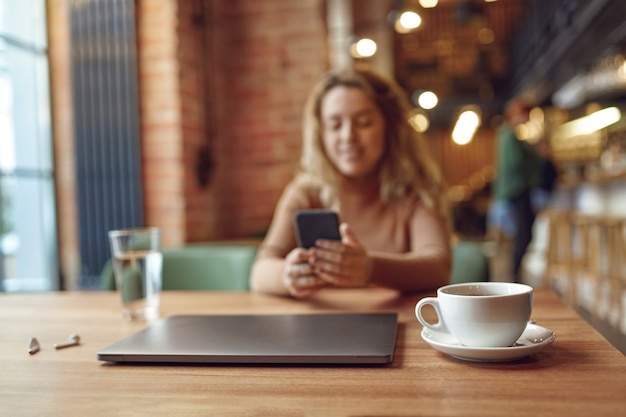 Blur of charming woman in stylish outfit surfing internet on modern smartphone while sitting at cafe